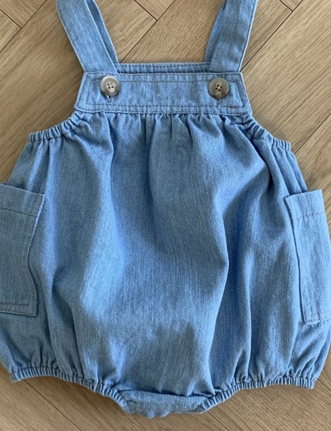 Ein kleiner, hellblauer Jeans-Strampler liegt flach auf einem Holzboden. Der ärmellose Bebe Holic Denim Pocket Bodysuit verfügt über verstellbare Träger mit Knöpfen, einen elastischen Bund und Beinöffnungen für Bewegungsfreiheit sowie zwei Seitentaschen.