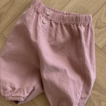 Ein Paar hellrosa Bebe Holic Natural Pants aus zerknittertem Stoff mit elastischem Bund und zwei Knöpfen vorne. Diese vielseitigen Must-have-Shorts haben lockere, geraffte Säume und werden flach auf einen Holzboden gelegt.