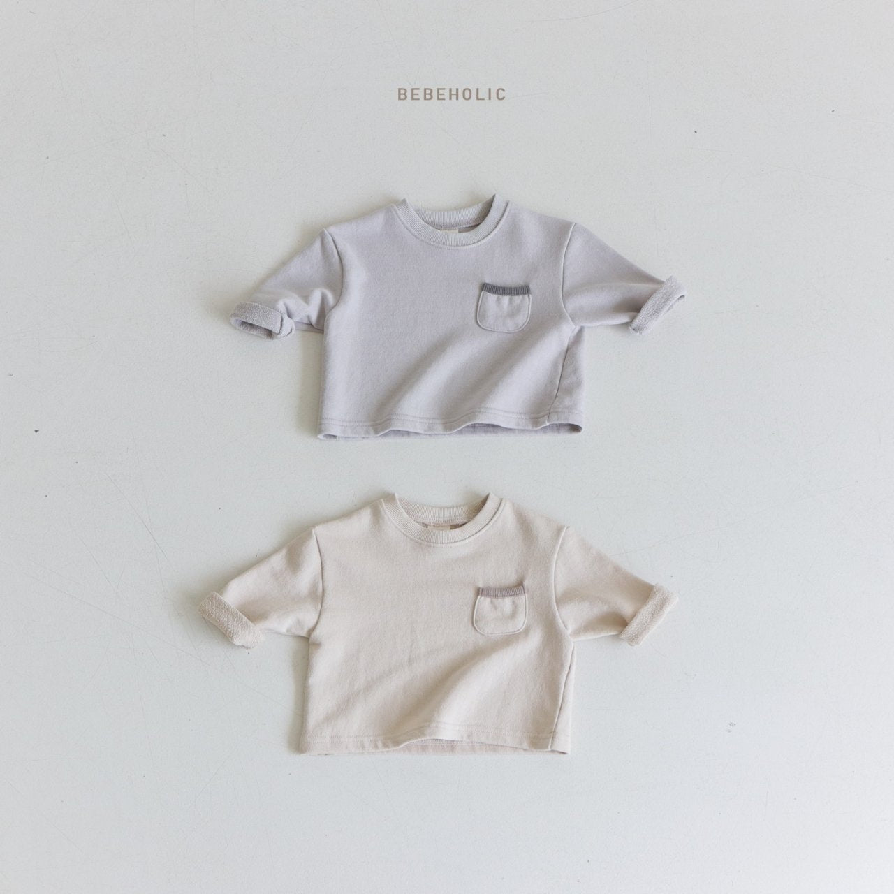 Auf neutralem Hintergrund sind zwei kleine, kurzärmelige Wiki-T-Shirts für Kleinkinder abgebildet. Das obere Shirt ist hellgrau und das untere Shirt ist beige. Jedes stylische Wiki-T-Shirt hat eine kleine Tasche auf der linken Brust und die Ärmel sind leicht gerollt. Über den Shirts steht der Markenname „Bebe Holic“.