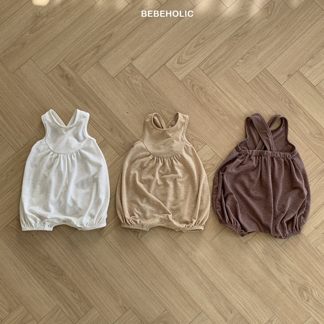 Drei Bebe Holic Leinen-Bodys mit Raffung liegen nebeneinander auf einem Holzboden. Diese ärmellosen Strampler in Weiß, Beige und Braun haben elastische Beinöffnungen für zusätzlichen Komfort für Kleinkinder.