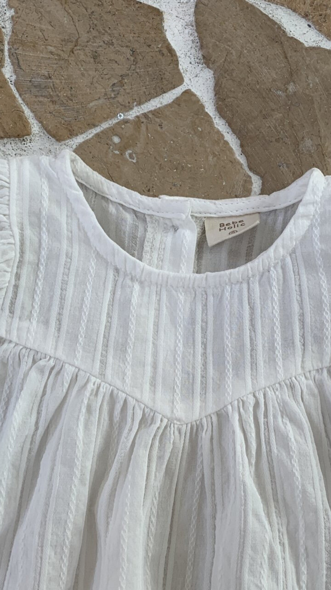 Nahaufnahme einer weißen, plissierten Raoul-Bluse mit Rüschendetails und einem Etikett mit der Marke „Bebe Holic“ auf einem strukturierten Hintergrund.