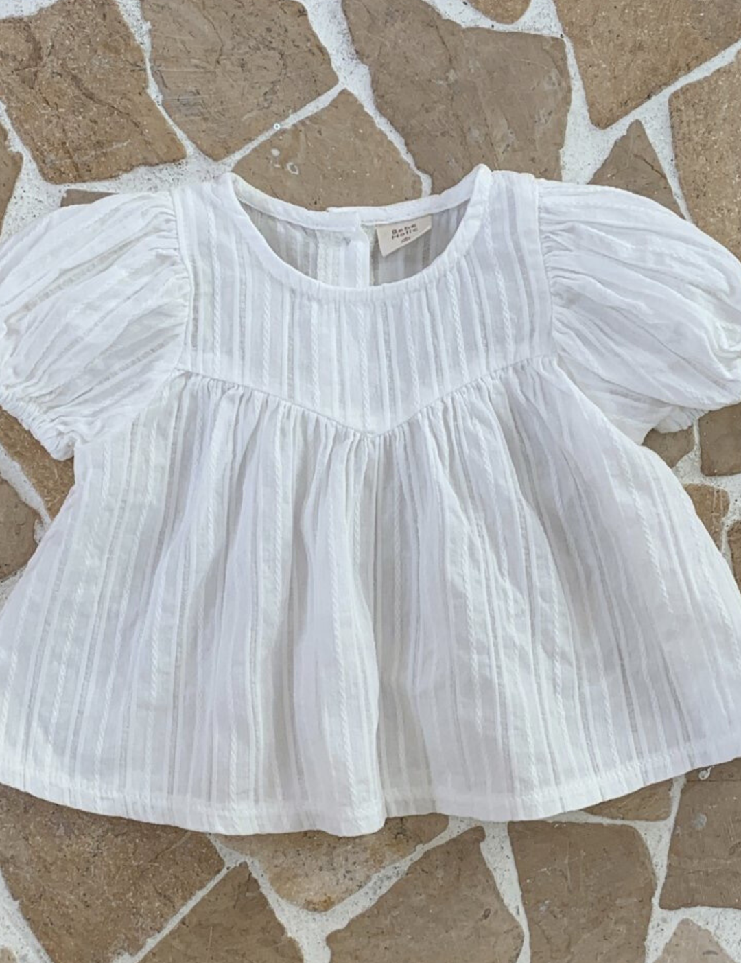 Eine weiße, kurzärmelige Bebe Holic Raoul Babybluse mit feinen Querstreifen und luftige Ärmelgestaltung, flach auf einem beige-braunen unregelmäßigen Fliesenboden liegend.