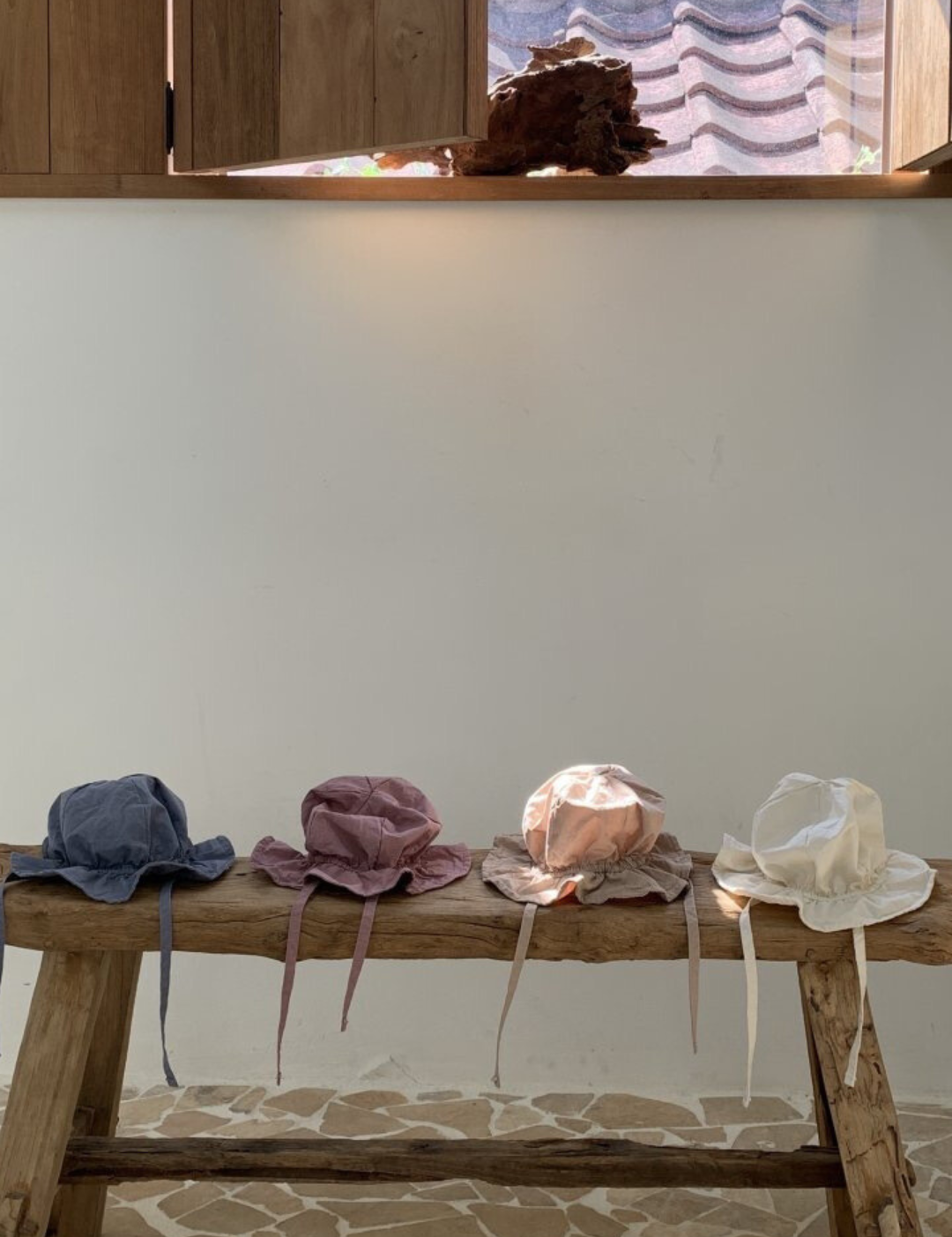 Eine Holzbank steht an einer cremefarbenen Wand und unter einem Fenster mit Holzläden. Vier Crunch Bucket Hats von Bebe Holic in Blau, Rosa, Beige und Weiß sind ordentlich auf der Bank angeordnet. Der Boden besteht aus unregelmäßig geformten Steinfliesen, die die natürliche Farbpalette des Raumes hervorheben.