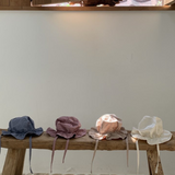 Eine Holzbank steht an einer cremefarbenen Wand und unter einem Fenster mit Holzläden. Vier Crunch Bucket Hats von Bebe Holic in Blau, Rosa, Beige und Weiß sind ordentlich auf der Bank angeordnet. Der Boden besteht aus unregelmäßig geformten Steinfliesen, die die natürliche Farbpalette des Raumes hervorheben.