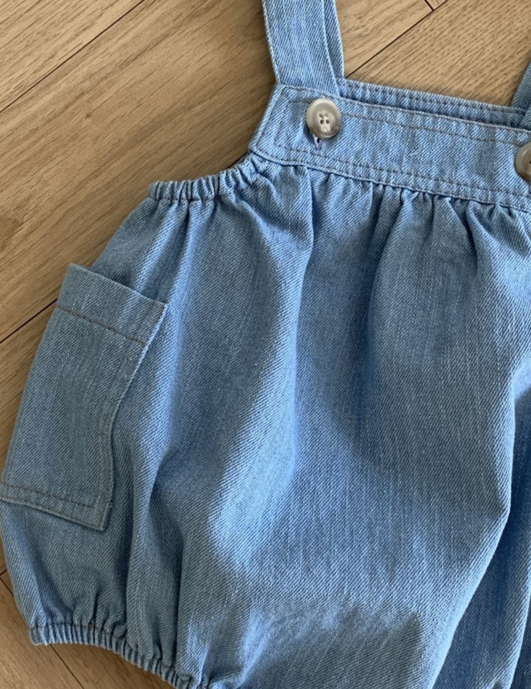 Ein Denim-Bodysuit mit Taschen von Bebe Holic verfügt über ein gerafftes Mieder, verstellbare Träger mit Knöpfen und einen elastischen Bund. Mit einer kleinen Tasche auf der linken Seite sorgt dieser Strampler für Bewegungsfreiheit für Ihren Kleinen. Er ist auf einem Holzboden ausgelegt.