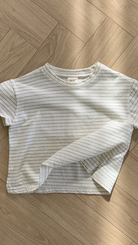 Ein weißes, kurzärmliges, kurzes T-Shirt mit dünnen schwarzen Querstreifen liegt ordentlich auf einem hölzernen Boden mit Fischgrätenmuster. Der Saum dieses Bebe Holic Basic ST Tee ist leicht angehoben und zeigt ein wenig den Stoff auf der Innenseite. Die Beleuchtung erzeugt sanfte Schatten auf dem Boden und unterstreicht seinen klassischen Stil.