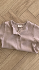 Ein weich aussehendes, kurzärmliges, geripptes Henley-Shirt in hellbeige Farbe liegt ordentlich gefaltet auf einem Holzparkettboden. Das Bebe Holic ST Tee besteht aus atmungsaktiver Baumwolle und hat einen geknöpften Ausschnitt mit einem offenen Knopf.