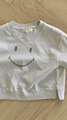 Ein hellgraues Sweatshirt aus weicher Baumwolle mit einem einfachen, großen Smiley-Design auf der Vorderseite wird ordentlich gefaltet und auf einen Holzboden gelegt. Auf dem Schild am Kragen steht „Smile Tee by Bebe Holic“.