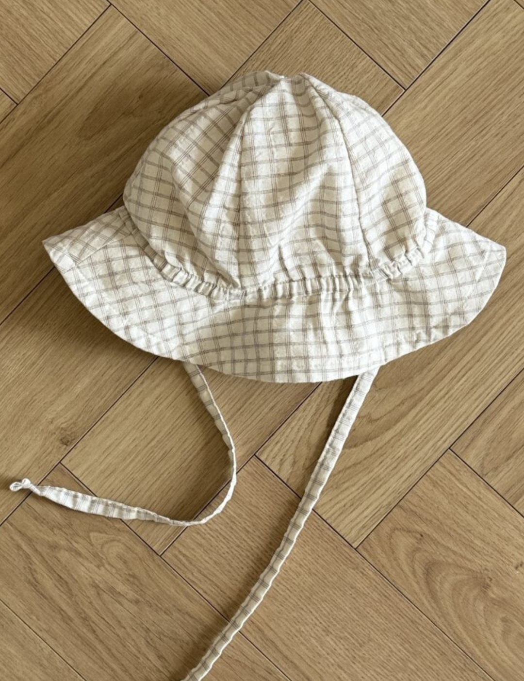 Ein weiß-beiger karierter Yogurt ST Bucket Hat von Bebe Holic, aus atmungsaktiven Materialien, mit breiter Krempe und Kinnriemen, liegt auf einem hellen Holzboden mit Fischgrätenmuster.