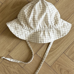 Ein weiß-beiger karierter Yogurt ST Bucket Hat von Bebe Holic, aus atmungsaktiven Materialien, mit breiter Krempe und Kinnriemen, liegt auf einem hellen Holzboden mit Fischgrätenmuster.