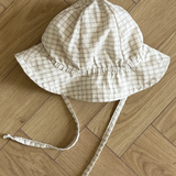 Ein weiß-beiger karierter Yogurt ST Bucket Hat von Bebe Holic, aus atmungsaktiven Materialien, mit breiter Krempe und Kinnriemen, liegt auf einem hellen Holzboden mit Fischgrätenmuster.