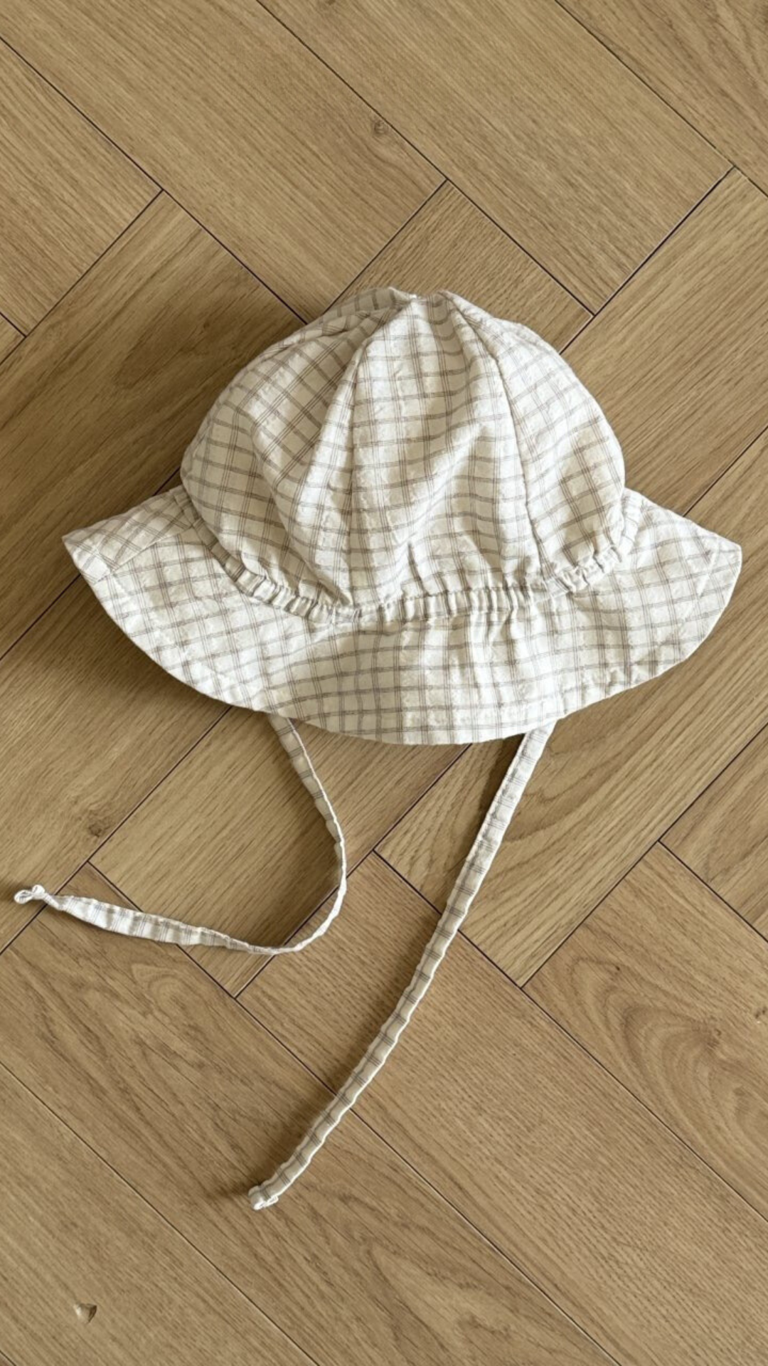 Ein weiß-beiger karierter Yogurt ST Bucket Hat von Bebe Holic, aus atmungsaktiven Materialien, mit breiter Krempe und Kinnriemen, liegt auf einem hellen Holzboden mit Fischgrätenmuster.