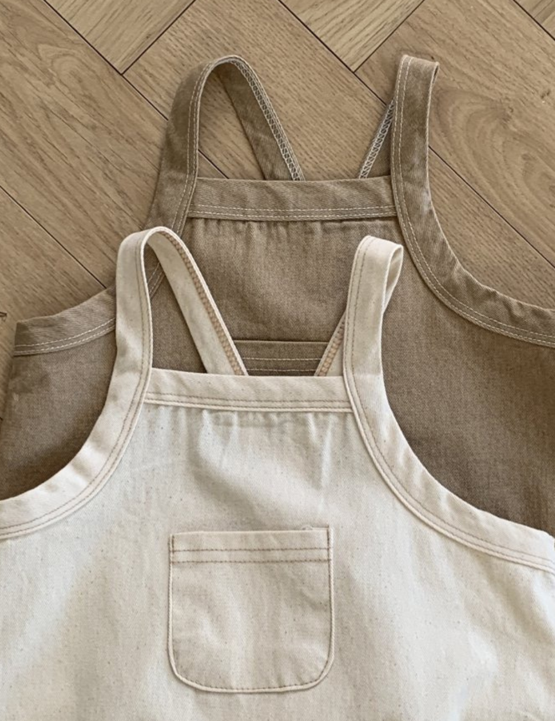 Zwei Bebeholic Twill Pocket Body Suits liegen flach auf einem Holzboden; einer ist taupe und der andere beige, beide mit Vordertaschen und Schulterriemen.