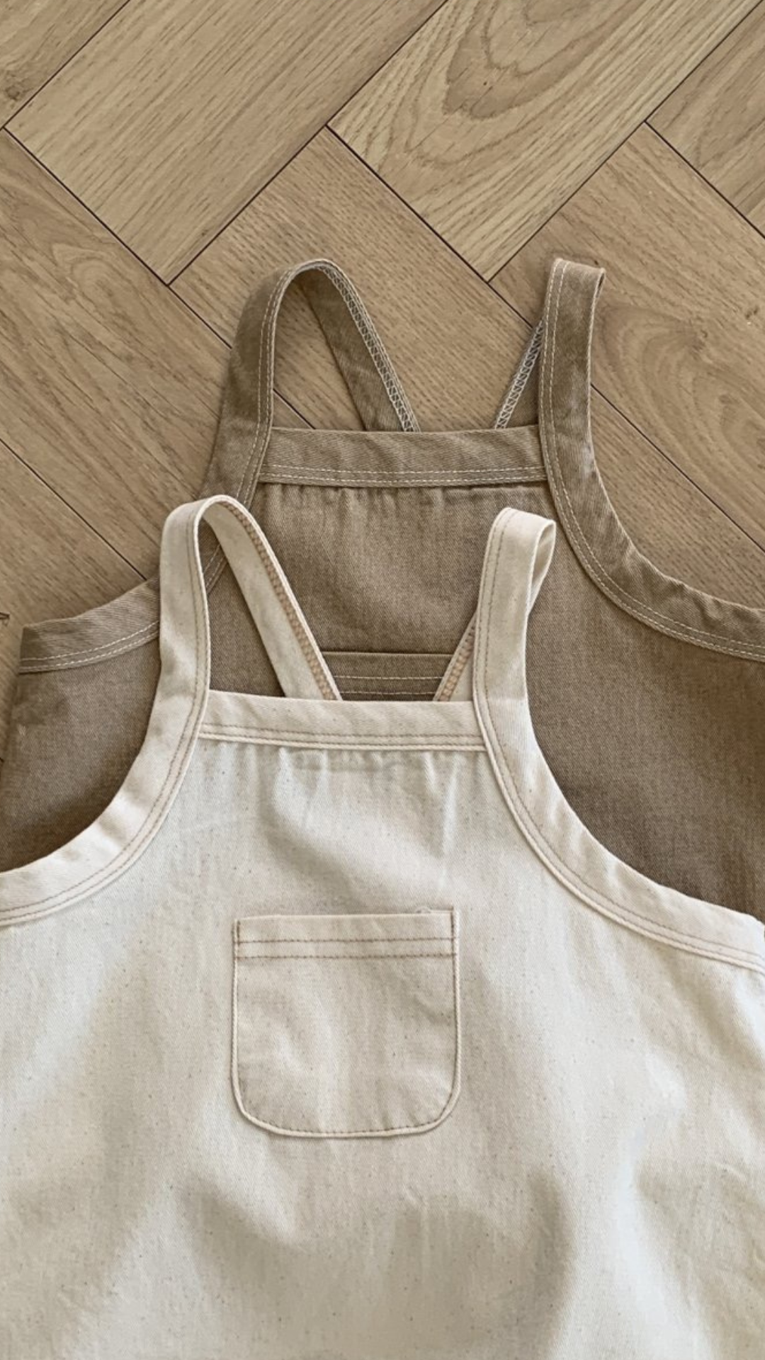 Zwei Bebeholic Twill Pocket Body Suits liegen flach auf einem Holzboden; einer ist taupe und der andere beige, beide mit Vordertaschen und Schulterriemen.