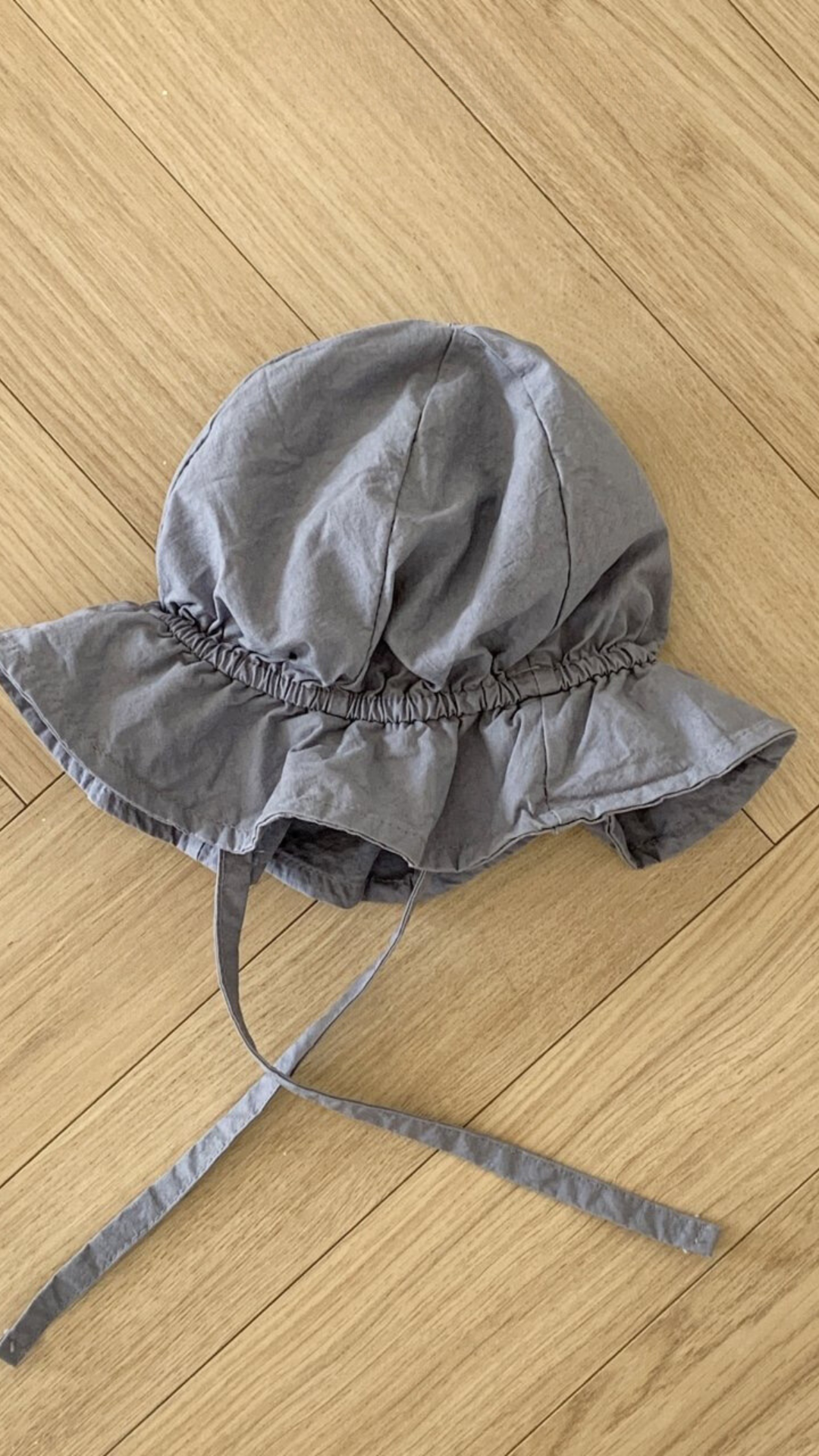 Ein grauer Crunch Bucket Hat aus der Bebe Holic-Kollektion mit breiter Krempe und Kinnriemen liegt auf einem Holzboden. Der Hut hat ein Gummiband um die Krone für einen gerafften Look, der seine natürliche Farbpalette perfekt ergänzt.