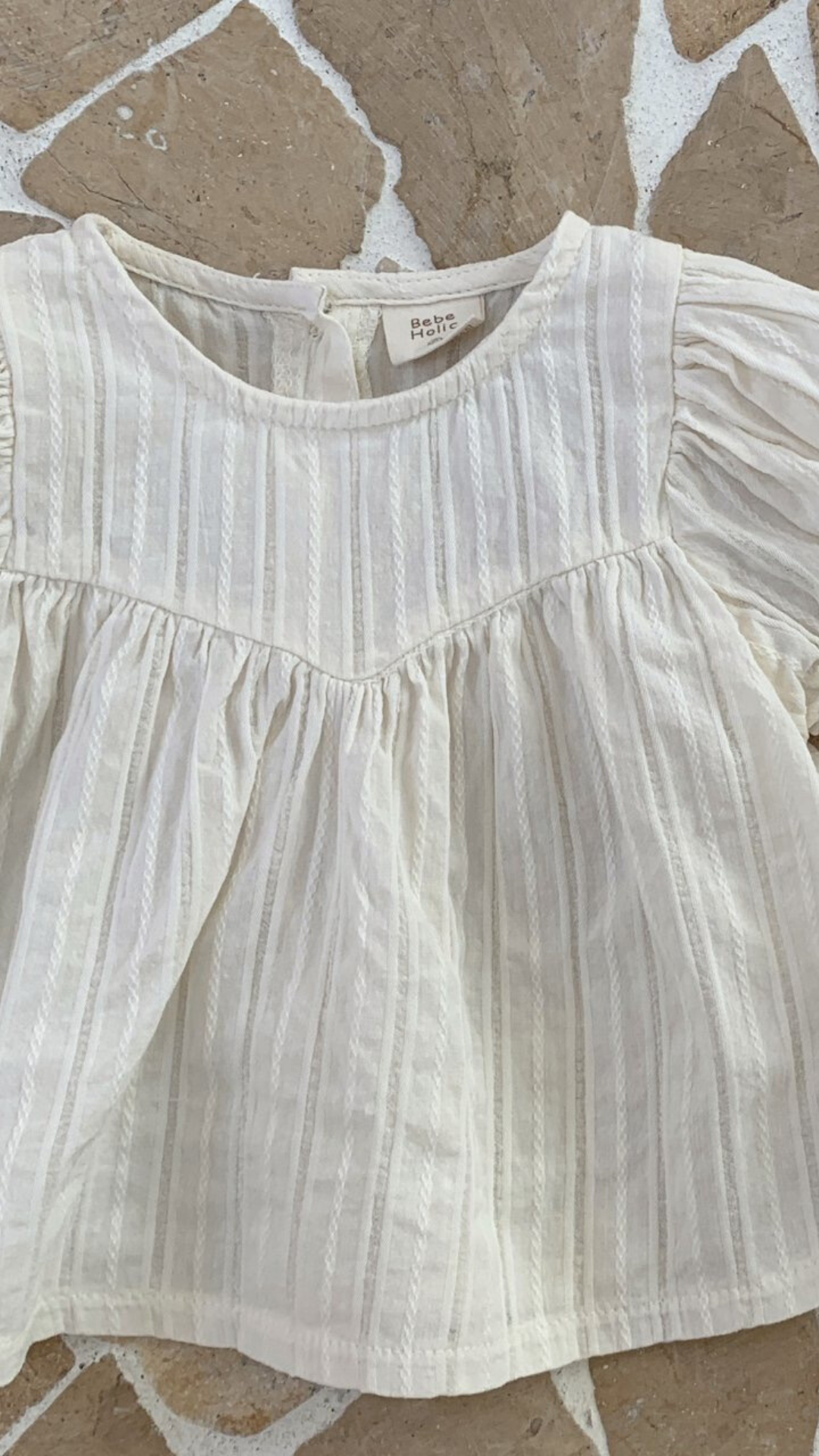 Eine Nahaufnahme einer weißen, strukturierten Raoul-Bluse von Bebe Holic mit Rüschendetails und kurzen Ärmeln, präsentiert auf einer gemusterten Oberfläche.