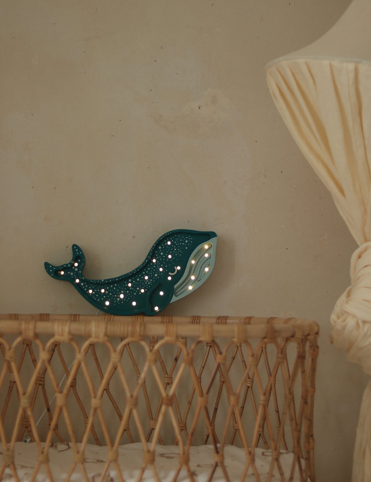 Eine Korbwiege mit weißer Matratze steht neben einer großen cremefarbenen Stehlampe mit Stoffschirm. Auf der Wiege steht eine Little Lights Whale Lamp | Galaxy Teal von Little Lights, die mit kleinen leuchtenden Punkten beleuchtet ist. Der Hintergrund zeigt ein Ozean-Abenteuer-Wandbild auf einer schlichten beigen Wand, perfekt für die Kinderzimmerdekoration.
