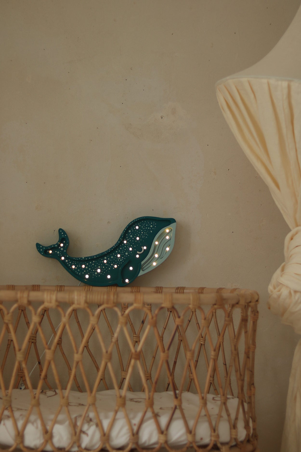 Eine Korbwiege mit weißer Matratze steht neben einer großen cremefarbenen Stehlampe mit Stoffschirm. Auf der Wiege steht eine Little Lights Whale Lamp | Galaxy Teal von Little Lights, die mit kleinen leuchtenden Punkten beleuchtet ist. Der Hintergrund zeigt ein Ozean-Abenteuer-Wandbild auf einer schlichten beigen Wand, perfekt für die Kinderzimmerdekoration.
