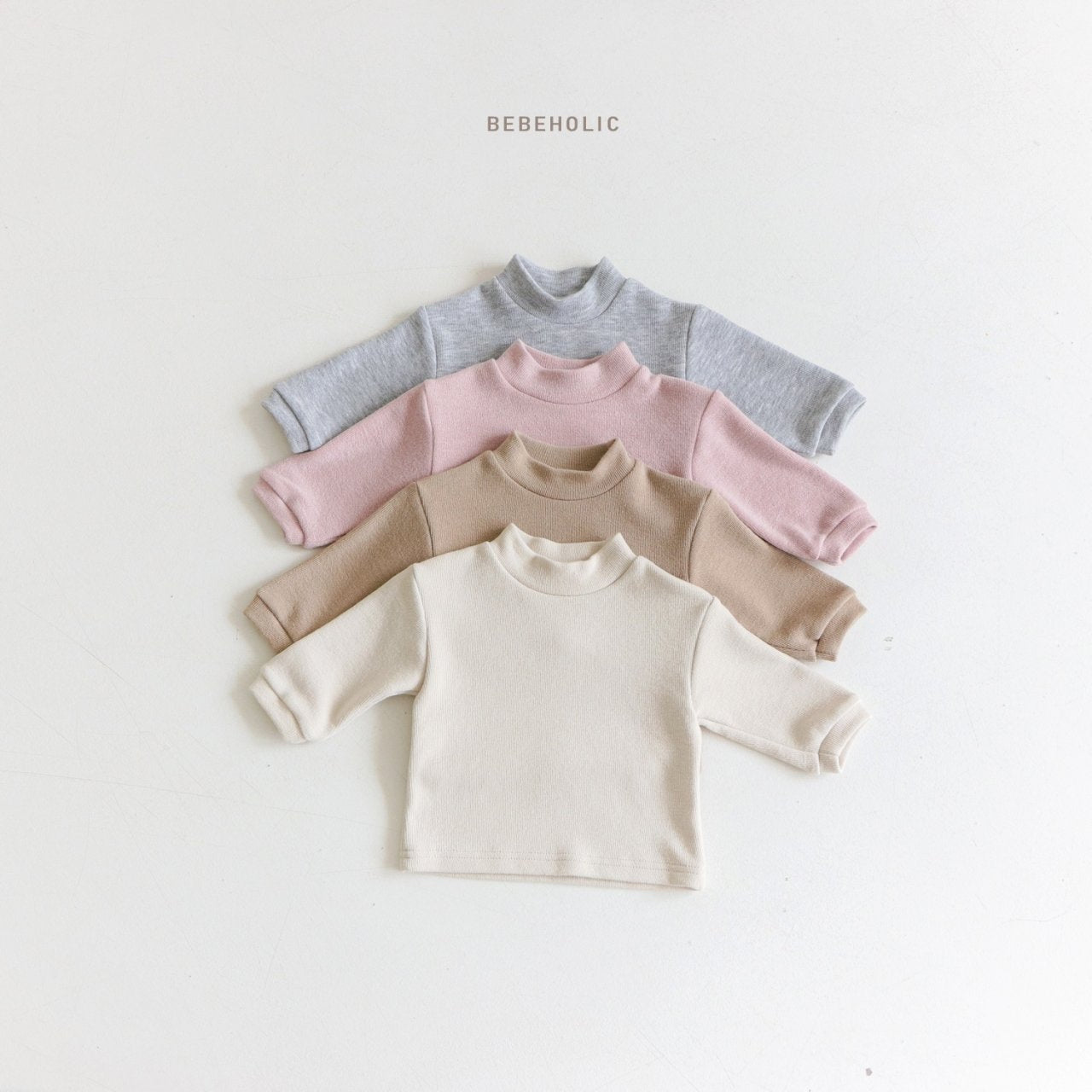 Vier langärmelige Babyshirts aus hautfreundlichem Material liegen ordentlich in einem vertikalen Stapel. Von oben nach unten sind die Shirts grau, rosa, beige und cremefarben. Über dem bequemen Tube Tee – Bequemes und Stilvolles T-Shirt für die Kleinsten-Kollektion ist auf weißem Hintergrund der Schriftzug „Bebe Holic“ aufgedruckt.