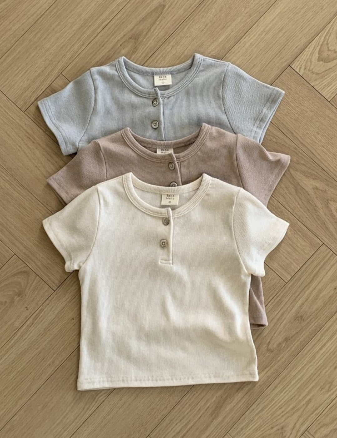 Drei Bebe Holic ST T-Shirts, ordentlich in einem vertikalen Stapel auf einem Holzboden angeordnet. Die Shirts aus atmungsaktiver Baumwolle sind in gedeckten Pastellfarben gehalten: oben hellblau, in der Mitte hellbraun und unten cremefarben. Jedes Shirt verfügt über ein kleines Knopfleisten-am-Kragen-Detail.