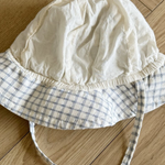 Eine weiße Babyhaube mit blau kariertem Rand und langen Bändern liegt auf einem Holzboden mit Fischgrätenmuster. Der Bebe Holic Yogurt ST Bucket Hat wirkt leicht zerknittert und hat eine weiche Stoffstruktur.