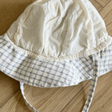 Eine weiße Babyhaube mit blau kariertem Rand und langen Bändern liegt auf einem Holzboden mit Fischgrätenmuster. Der Bebe Holic Yogurt ST Bucket Hat wirkt leicht zerknittert und hat eine weiche Stoffstruktur.