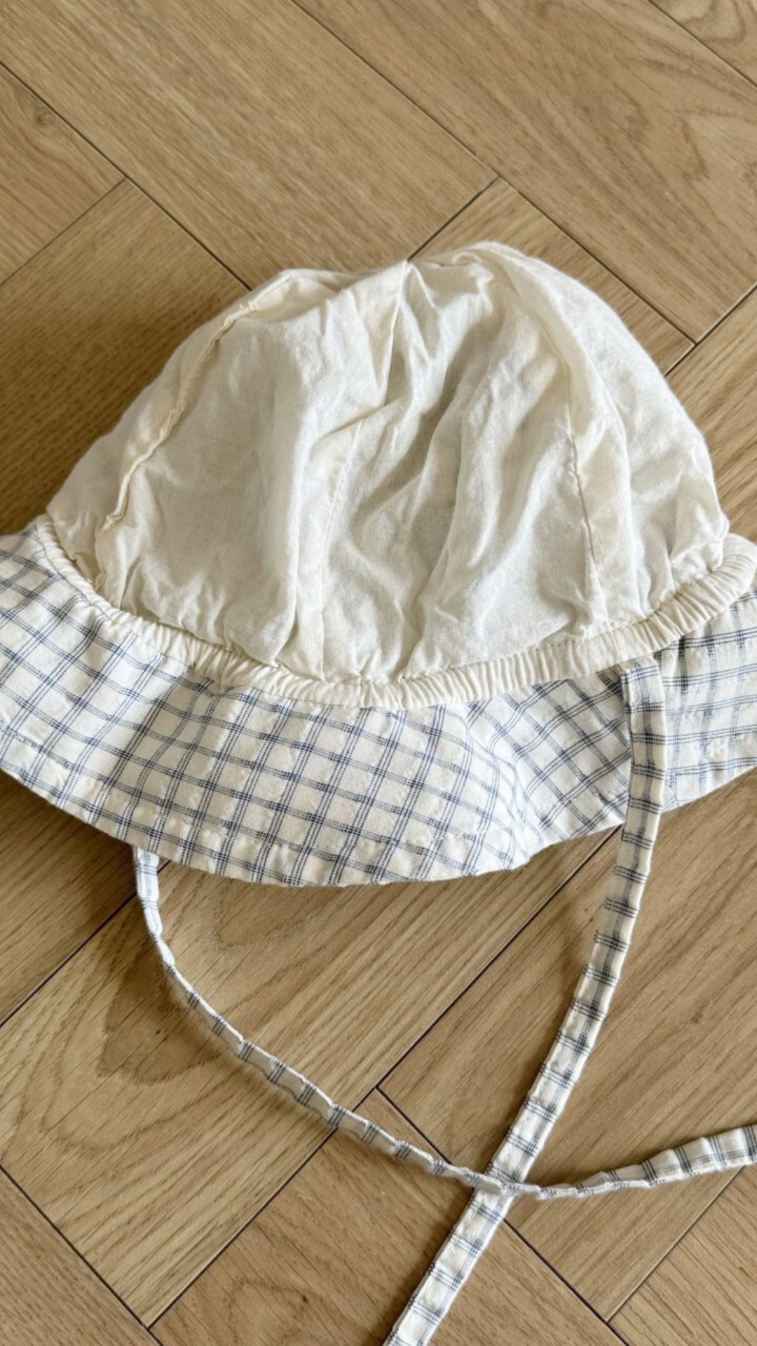 Eine weiße Babyhaube mit blau kariertem Rand und langen Bändern liegt auf einem Holzboden mit Fischgrätenmuster. Der Bebe Holic Yogurt ST Bucket Hat wirkt leicht zerknittert und hat eine weiche Stoffstruktur.