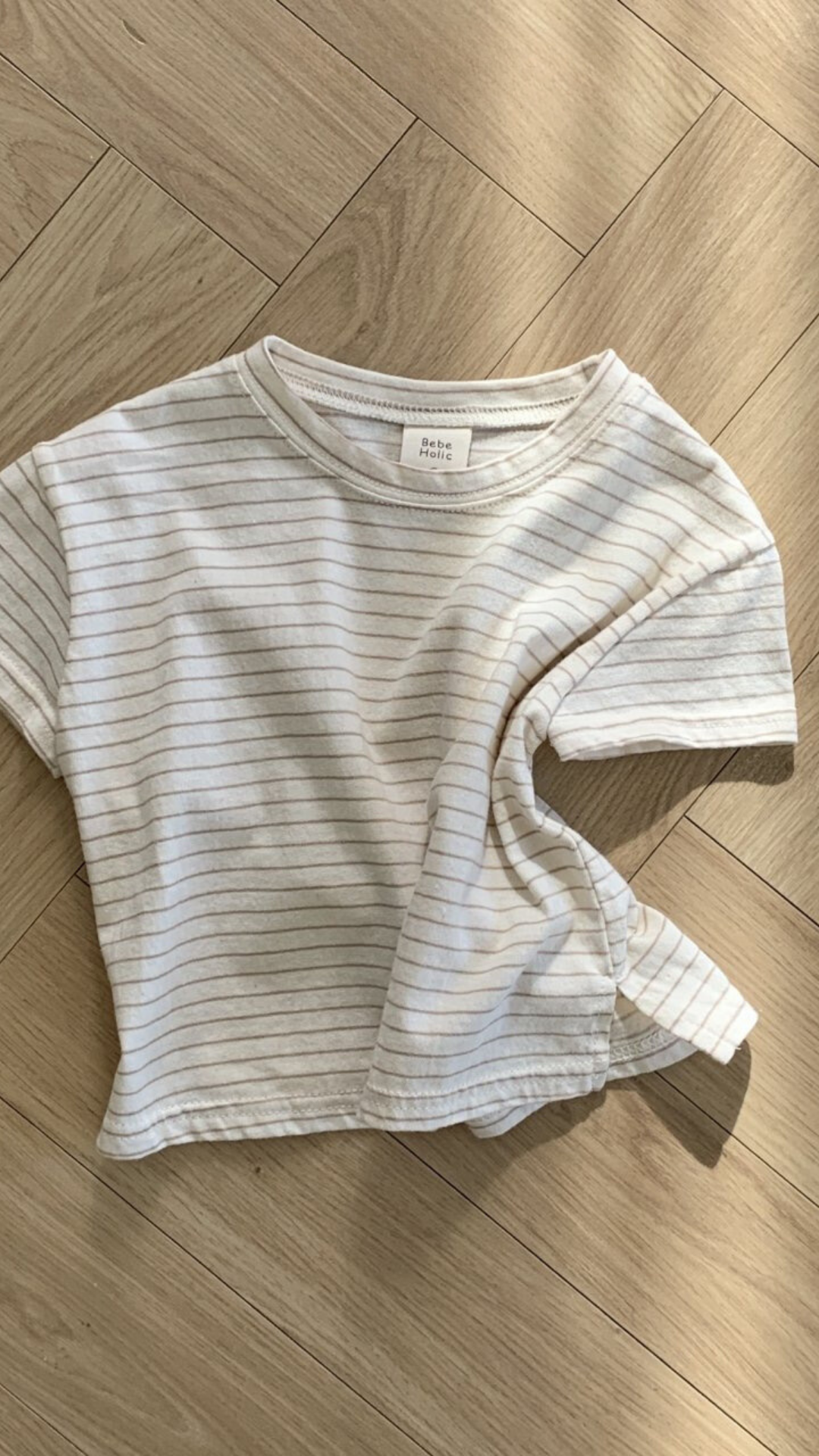 Ein weißes kurzärmliges Basic ST Tee mit dünnen schwarzen Querstreifen von Bebe Holic liegt ordentlich auf einem Holzboden mit Fischgrätenmuster und zeigt seinen klassischen Stil. Der Stoff des T-Shirts ist leicht zerknittert.