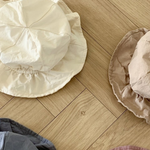 Vier zerknitterte Crunch Bucket Hats von Bebe Holic in einer natürlichen Farbpalette aus Weiß, Beige, Blau und Kastanienbraun sind auf einem hölzernen Boden mit Fischgrätenmuster ausgebreitet. Die Hüte scheinen aus einem leichten, lässigen Stoff gefertigt zu sein.