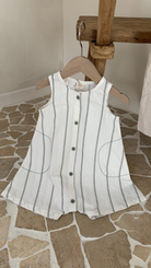 Ein Bebe Holic Daisy ST Bodysuit mit elegantem Streifendesign hängt auf einem hölzernen Kleiderbügel. Das Oberteil hat unten eine ausgestellte Form und wird auf einem Boden mit Steinmuster und Holzmöbeln im Hintergrund präsentiert, was einen modischeren Look ergibt.