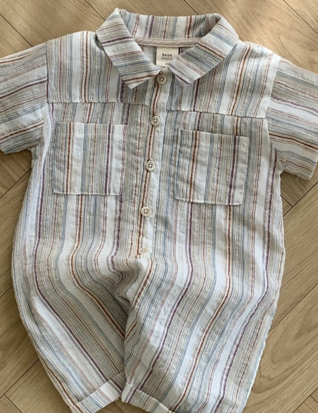 Ein kurzärmeliger Aurora-Bodysuit von Bebe Holic liegt auf einem Holzboden. Der Strampler, der an gestreifte Hemden erinnert, hat einen Kragen, zwei Brusttaschen und Knöpfe auf der Vorderseite. Er besticht durch ein Streifenmuster in verschiedenen Blau-, Rot- und Beigetönen.