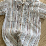 Ein kurzärmeliger Aurora-Bodysuit von Bebe Holic liegt auf einem Holzboden. Der Strampler, der an gestreifte Hemden erinnert, hat einen Kragen, zwei Brusttaschen und Knöpfe auf der Vorderseite. Er besticht durch ein Streifenmuster in verschiedenen Blau-, Rot- und Beigetönen.