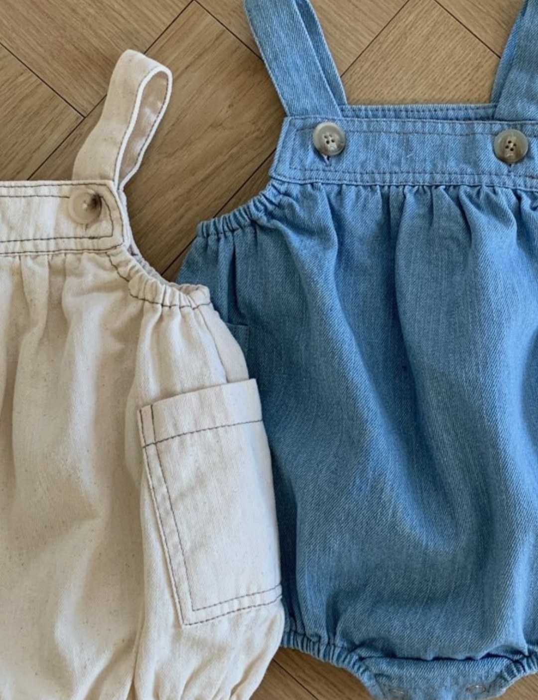 Zwei Baby-Strampelanzug-Outfits auf Holzoberfläche: einer ist beige mit Tasche und geknöpften Trägern, der andere ist aus blauem Denim mit verstellbaren Trägern. Beide Bebe Holic Denim Pocket Body Suits haben ein gerafftes, bauschiges Design für Bewegungsfreiheit.