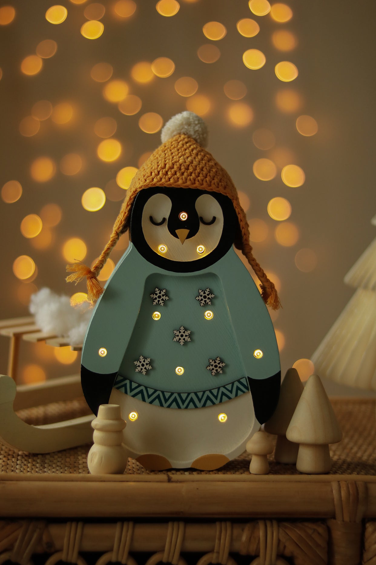 Festliche Little Lights – Baby Penguin Mini Christmas Edition | Blau mit leuchtenden Lichtern trägt eine senfgelbe Strickmütze und einen blauen Pullover mit Schneeflocken. Er steht auf einer Holzoberfläche mit verschwommenen Bokeh-Lichtern im Hintergrund und schafft eine warme und gemütliche Atmosphäre, perfekt als Kinderzimmer-Dekoration.