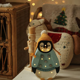 Eine gemütliche Weihnachtsszene mit einer dekorativen Pinguinfigur, die eine gelbe Mütze und einen blauen Pullover mit Schneeflockenmotiven trägt. Die Little Lights – Baby Penguin Mini Christmas Edition | Blue strahlt ein warmes Licht aus, perfekt für die Kinderzimmer-Dekoration, und im Hintergrund befinden sich festliche Kissen auf einem Korbstuhl mit einem Weihnachtsbaummotiv.