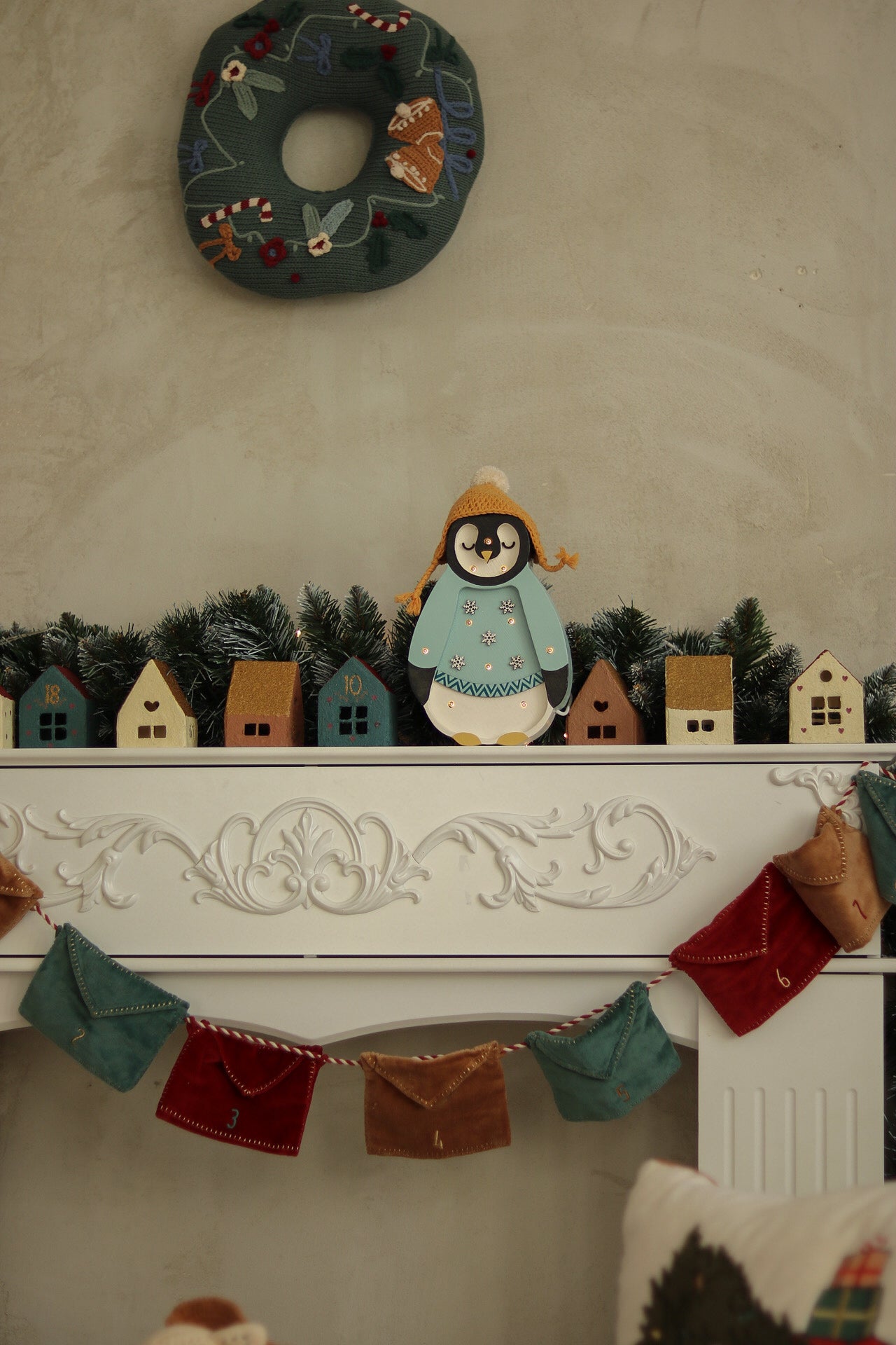 Auf einem festlichen Kaminsims steht ein Little Lights - Baby Penguin Mini Christmas Edition | Blue von Little Lights mit Wintermantel und Mütze zwischen Miniatur-Holzhäusern und grünen Tannenzweigen. Darüber hängt ein Weihnachtskranz. Darunter hängen bunte Stoffwimpel an der Vorderseite des Kaminsimses und bilden die perfekte Kinderzimmer-Dekoration.