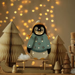 A Little Lights – Baby Penguin Mini Christmas Edition | Blue steht auf einem kleinen Schlitten. Er ist umgeben von Papierbäumen und weiteren Holzdekorationen, eine ideale Kinderzimmer-Dekoration. Warme Lichterketten erzeugen im Hintergrund einen Bokeh-Effekt.