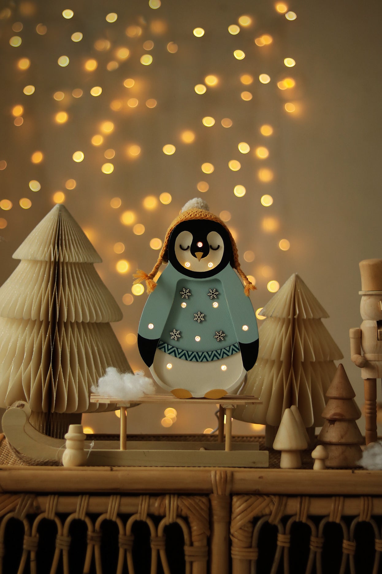 A Little Lights – Baby Penguin Mini Christmas Edition | Blue steht auf einem kleinen Schlitten. Er ist umgeben von Papierbäumen und weiteren Holzdekorationen, eine ideale Kinderzimmer-Dekoration. Warme Lichterketten erzeugen im Hintergrund einen Bokeh-Effekt.