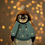 Ein dekorativer Little Lights - Baby Penguin Mini Christmas Edition | Blue von Little Lights mit einer gemütlichen gelben Strickmütze und einem blauen Pullover, der mit Schneeflocken geschmückt ist, steht auf einer Korboberfläche. Im Hintergrund leuchten warmweiße Lichterketten und schaffen eine festliche Atmosphäre, die perfekt zur Kinderzimmer-Dekoration passt.