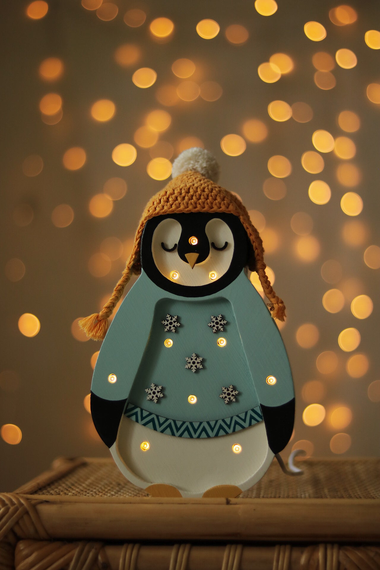 Ein dekorativer Little Lights - Baby Penguin Mini Christmas Edition | Blue von Little Lights mit einer gemütlichen gelben Strickmütze und einem blauen Pullover, der mit Schneeflocken geschmückt ist, steht auf einer Korboberfläche. Im Hintergrund leuchten warmweiße Lichterketten und schaffen eine festliche Atmosphäre, die perfekt zur Kinderzimmer-Dekoration passt.