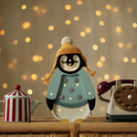 Eine festliche Szene zeigt eine süße Pinguinfigur, die eine gelbe Strickmütze mit Bommel und einen hellblauen Mantel mit Schneeflocken trägt. Der Pinguin, der gleichzeitig ein entzückender Little Lights - Baby Penguin Mini Christmas Edition | Blue von Little Lights ist, ist von einer rot-weiß gestreiften Teekanne und einem Vintage-Wählscheibentelefon auf einer Korboberfläche umgeben, mit funkelnden Lichtern im Hintergrund.