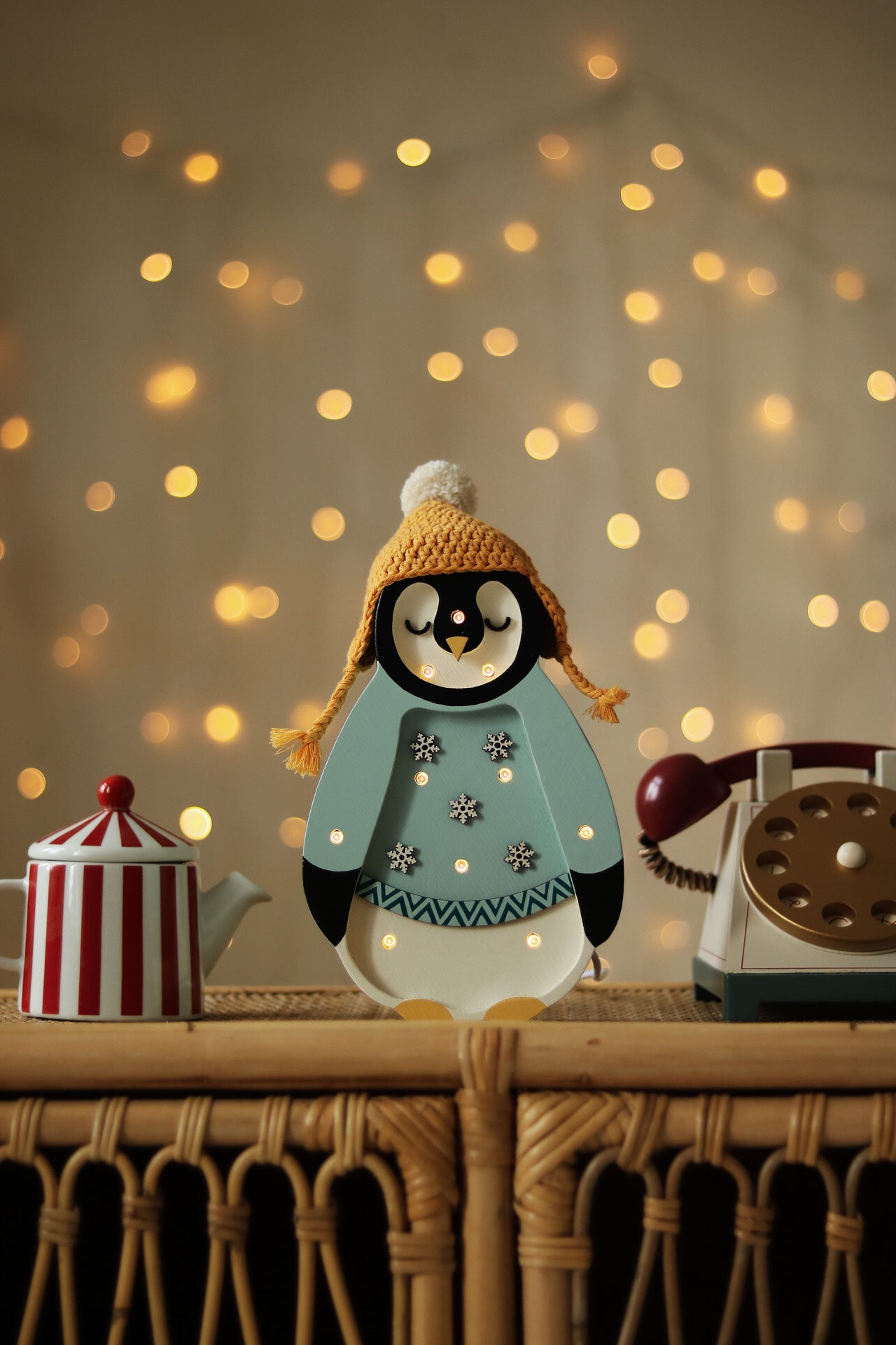 Eine festliche Szene zeigt eine süße Pinguinfigur, die eine gelbe Strickmütze mit Bommel und einen hellblauen Mantel mit Schneeflocken trägt. Der Pinguin, der gleichzeitig ein entzückender Little Lights - Baby Penguin Mini Christmas Edition | Blue von Little Lights ist, ist von einer rot-weiß gestreiften Teekanne und einem Vintage-Wählscheibentelefon auf einer Korboberfläche umgeben, mit funkelnden Lichtern im Hintergrund.