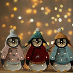 Drei kleine Lichter – Baby-Pinguin Mini-Weihnachtsedition | Blaue Figuren mit festlichen Hüten und Pullovern stehen auf einer Korboberfläche. In jedem Pinguin sind kleine leuchtende Lichter in den Körper eingearbeitet, perfekt als Nachtlicht für die Kinderzimmer-Dekoration. Der Hintergrund weist einen warmen Bokeh-Effekt aus kreisförmigen Lichtpunkten auf, der eine gemütliche, festliche Atmosphäre schafft.