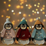 Drei kleine Lichter – Baby-Pinguin Mini-Weihnachtsedition | Blaue Figuren mit festlichen Hüten und Pullovern stehen auf einer Korboberfläche. In jedem Pinguin sind kleine leuchtende Lichter in den Körper eingearbeitet, perfekt als Nachtlicht für die Kinderzimmer-Dekoration. Der Hintergrund weist einen warmen Bokeh-Effekt aus kreisförmigen Lichtpunkten auf, der eine gemütliche, festliche Atmosphäre schafft.