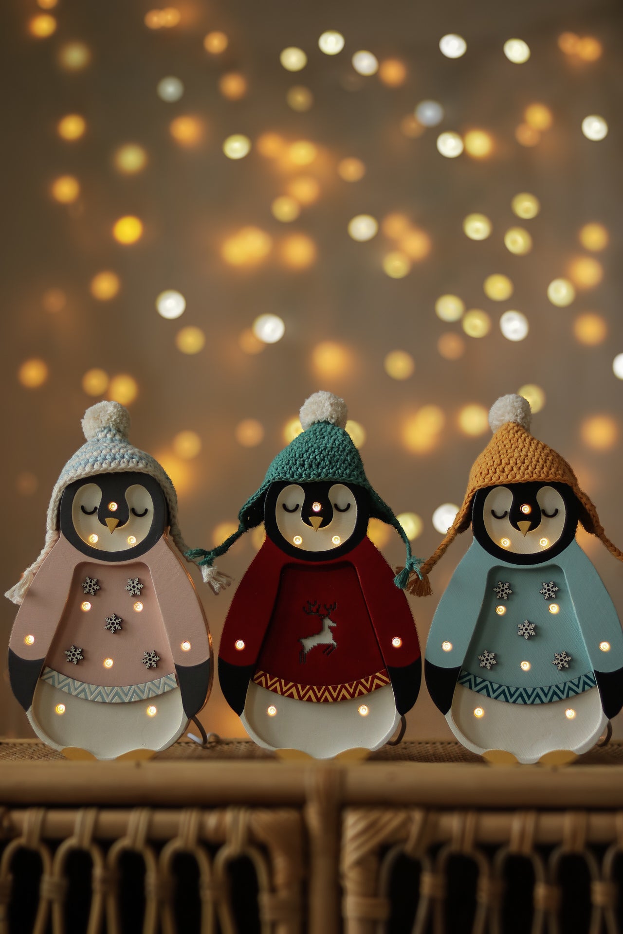 Drei kleine Lichter – Baby-Pinguin Mini-Weihnachtsedition | Blaue Figuren mit festlichen Hüten und Pullovern stehen auf einer Korboberfläche. In jedem Pinguin sind kleine leuchtende Lichter in den Körper eingearbeitet, perfekt als Nachtlicht für die Kinderzimmer-Dekoration. Der Hintergrund weist einen warmen Bokeh-Effekt aus kreisförmigen Lichtpunkten auf, der eine gemütliche, festliche Atmosphäre schafft.