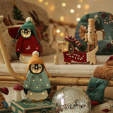 Eine gemütliche Weihnachtsszene zeigt zwei Pinguinfiguren in Winterkleidung und eine Dackelfigur mit Strickmütze. Ein Nussknacker, eine Discokugel und festliche Dekorationen sind überall verteilt. Weiches, warmes Licht von Little Lights – Baby Penguin Mini Christmas Edition | Blue von Little Lights und weihnachtlich gestaltete Kissen sorgen für eine gemütliche Atmosphäre.