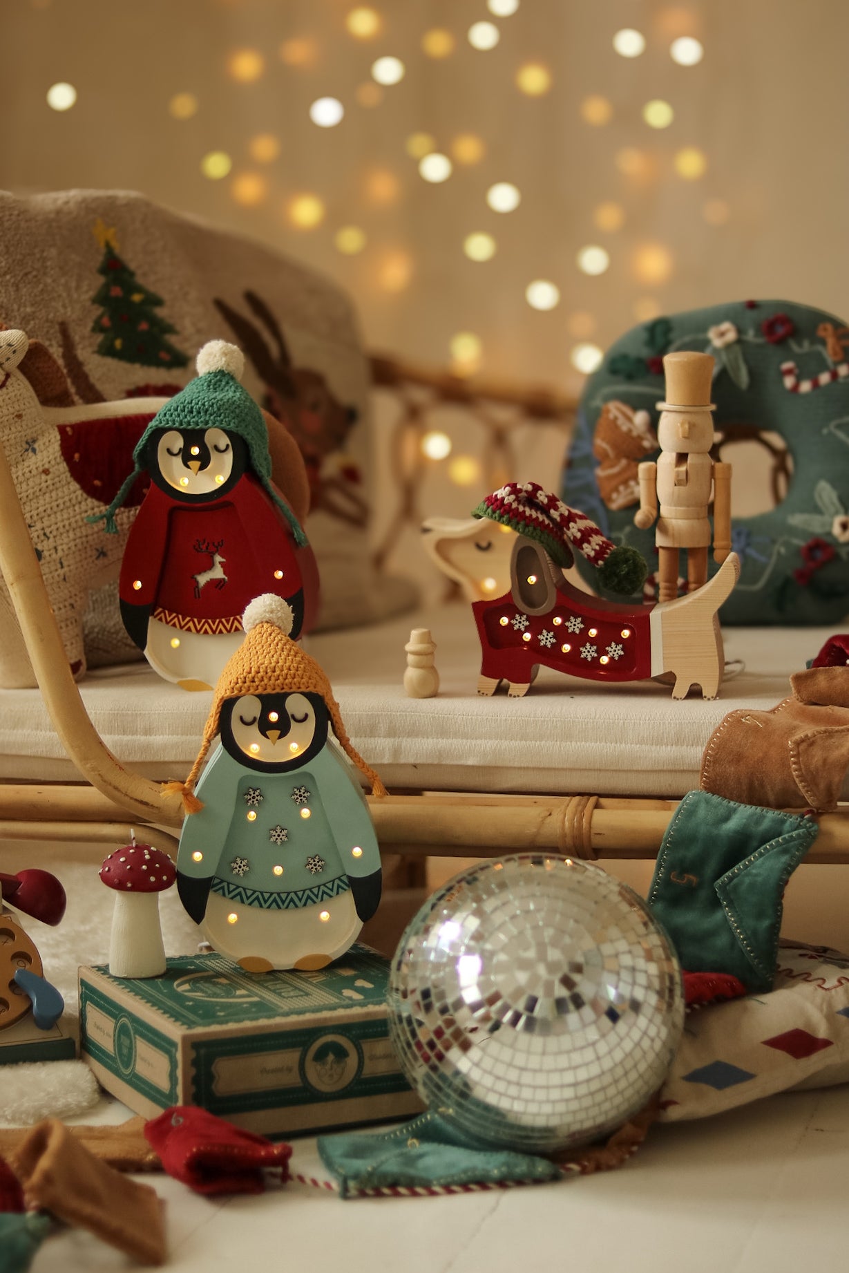 Eine gemütliche Weihnachtsszene zeigt zwei Pinguinfiguren in Winterkleidung und eine Dackelfigur mit Strickmütze. Ein Nussknacker, eine Discokugel und festliche Dekorationen sind überall verteilt. Weiches, warmes Licht von Little Lights – Baby Penguin Mini Christmas Edition | Blue von Little Lights und weihnachtlich gestaltete Kissen sorgen für eine gemütliche Atmosphäre.