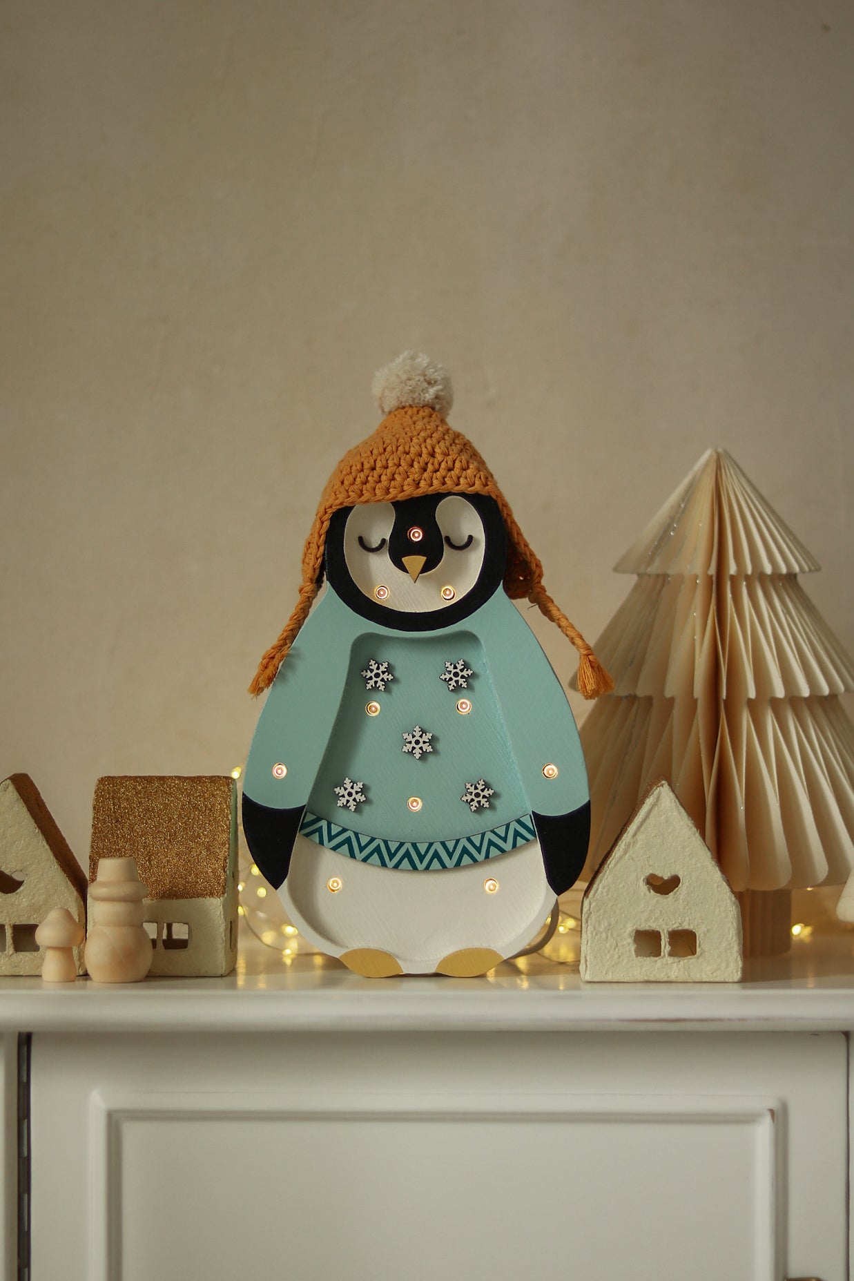 Eine dekorative Pinguinfigur mit einer orangefarbenen Strickmütze und einem hellblauen Pullover ist von kleinen Holzhäusern und einem Baum umgeben. Die kleinen Lichter – Baby Penguin Mini Christmas Edition | Blau und die Häuser werden von kleinen Lichtern beleuchtet und schaffen eine gemütliche Kinderzimmer-Dekoration mit Weihnachtsszene auf einer weißen Oberfläche.