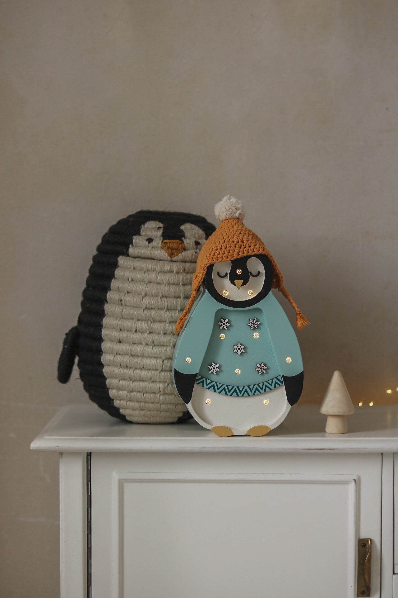 Eine bezaubernde Kinderzimmer-Dekoration mit zwei Pinguinfiguren: einem schwarz-weißen Plüschpinguin und einem kleineren hölzernen Pinguin mit Häkelmütze und hellblauem Pullover mit Schneeflockenmotiv. Sie stehen auf einem weißen Schrank neben einem kleinen Holzbaum. Die Szene enthält die Little Lights - Baby Penguin Mini Christmas Edition | Blue von Little Lights.