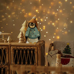 Eine festliche Szene mit warmen Weihnachtslichtern im Hintergrund. Im Vordergrund ein Korbschrank, dekoriert mit einem Little Lights – Baby Penguin Mini Christmas Edition | Blau von Little Lights und einem Miniatur-Weihnachtsbaum aus Holz. Neben dem Schrank steht ein gemütlicher Stuhl mit weihnachtlichen Kissen und sorgt für die perfekte Kinderzimmer-Dekoration.